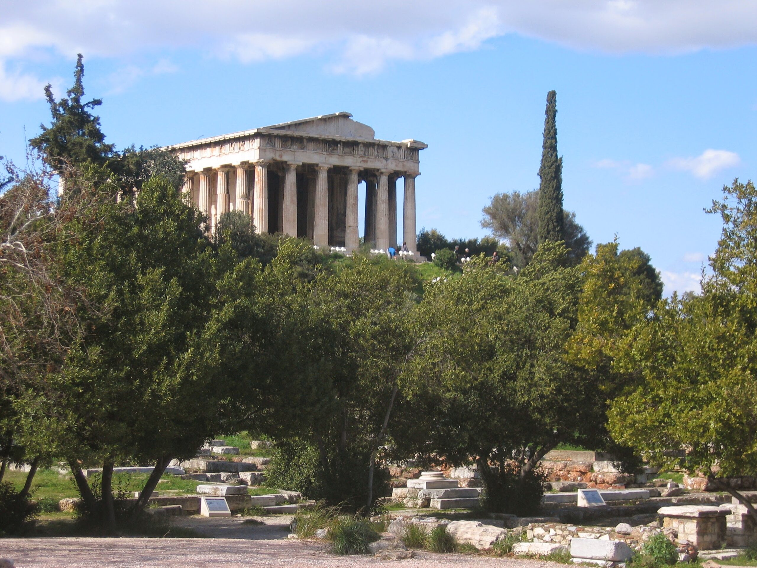 The Ancient Agora of Athens seen from below, with the foundations and cut blocks of the public buildings among olive trees and the complete Temple of Hephaestus on the hill above