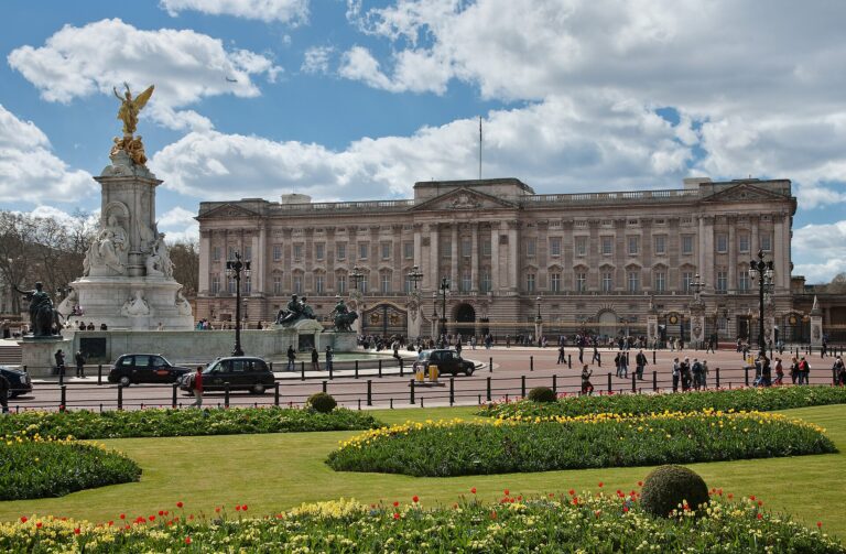 The East Front of Buckingham Palace in spring sunshine, with its railings and gates, the Victoria Memorial topped by a gilded figure on the left and tulip beds in the foreground