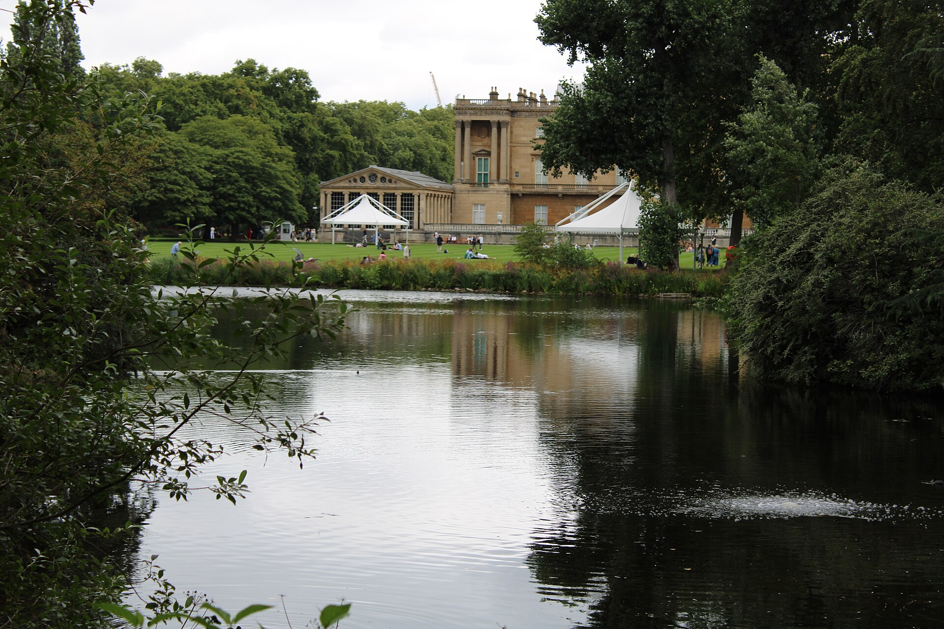 Buckingham Palace seen across the lake in its private garden, with summer visitors sitting on the lawn and white canopies in front of the building