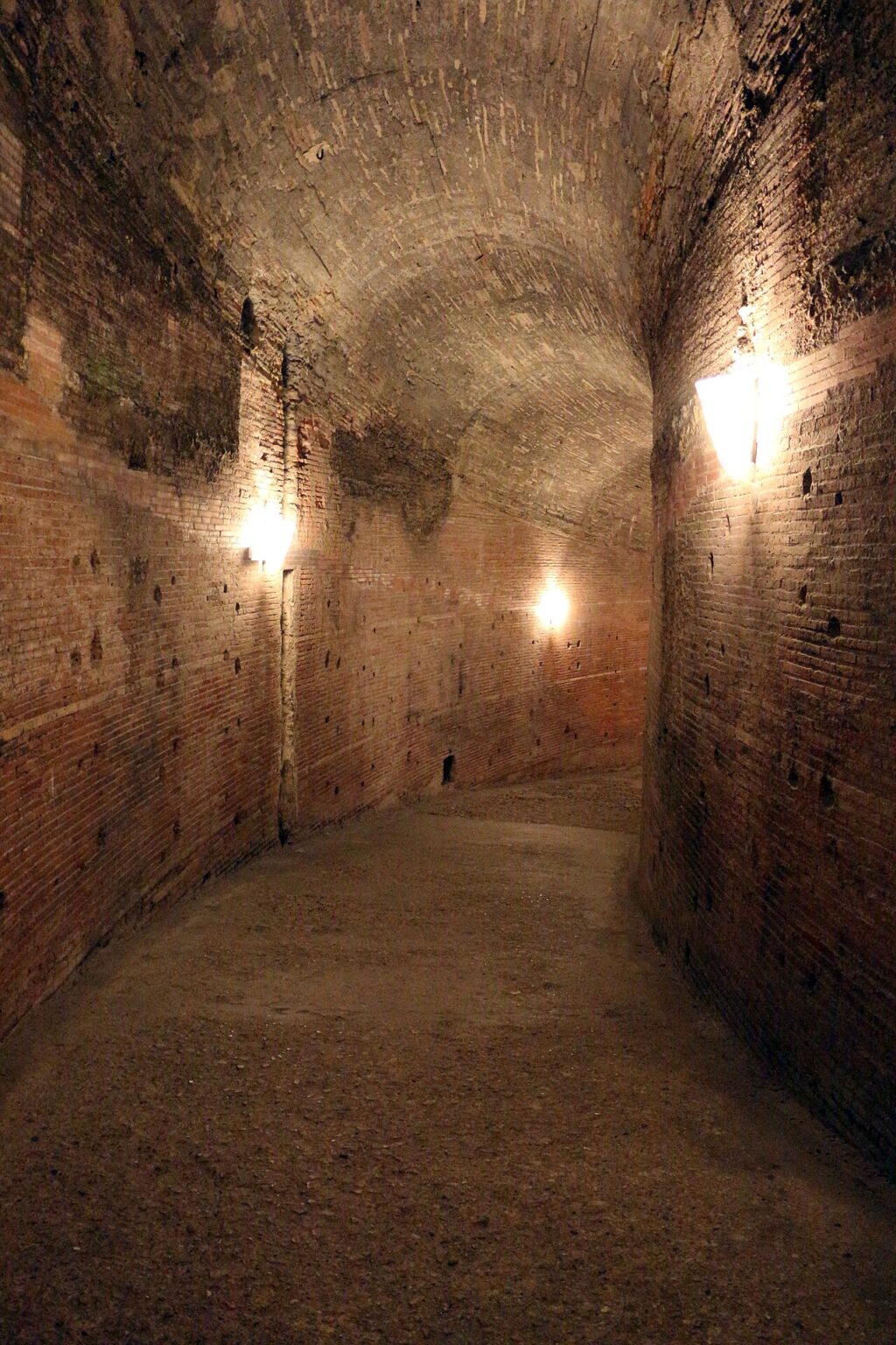 The dim brick-vaulted helical ramp inside Castel Sant'Angelo, lit by wall lamps, curving upwards