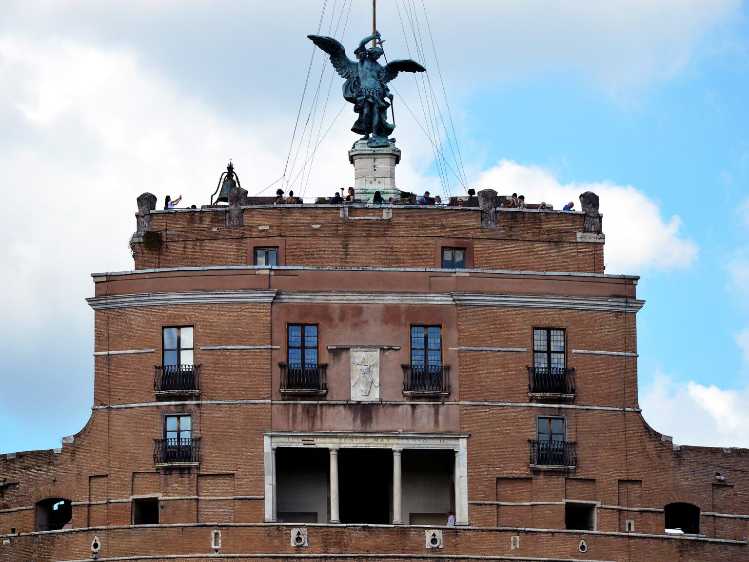 The top of Castel Sant'Angelo against a blue sky, with visitors on the terrace below the bronze statue of the angel