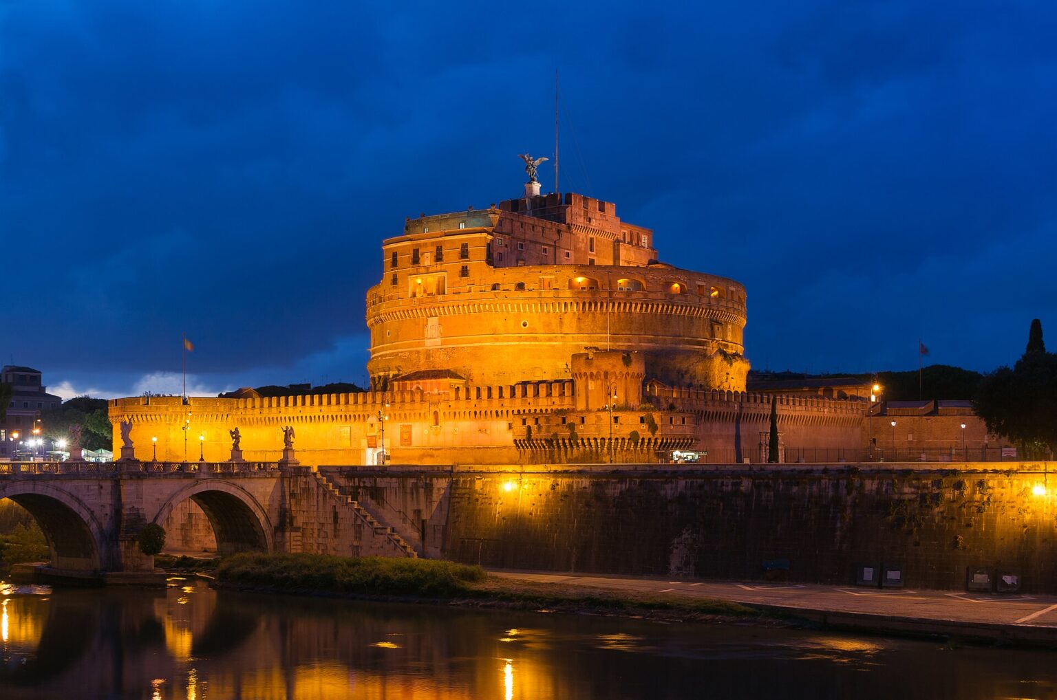 Castel Sant'Angelo lit up at dusk above the Tiber, with the Ponte Sant'Angelo and its reflection in the river in front