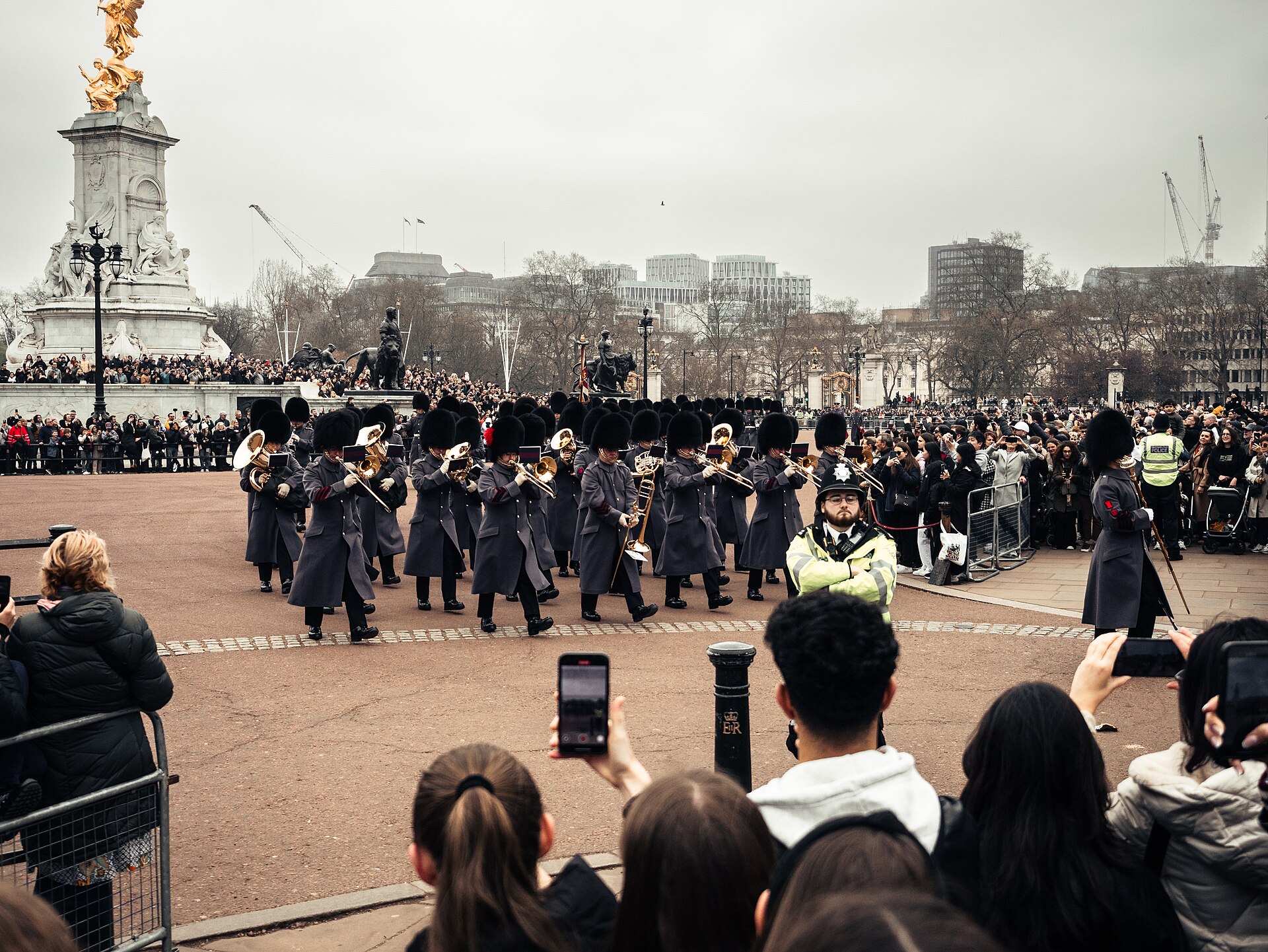A military band in grey greatcoats and bearskin hats marching past the Victoria Memorial during the Changing the Guard, watched by crowds behind barriers holding up their phones