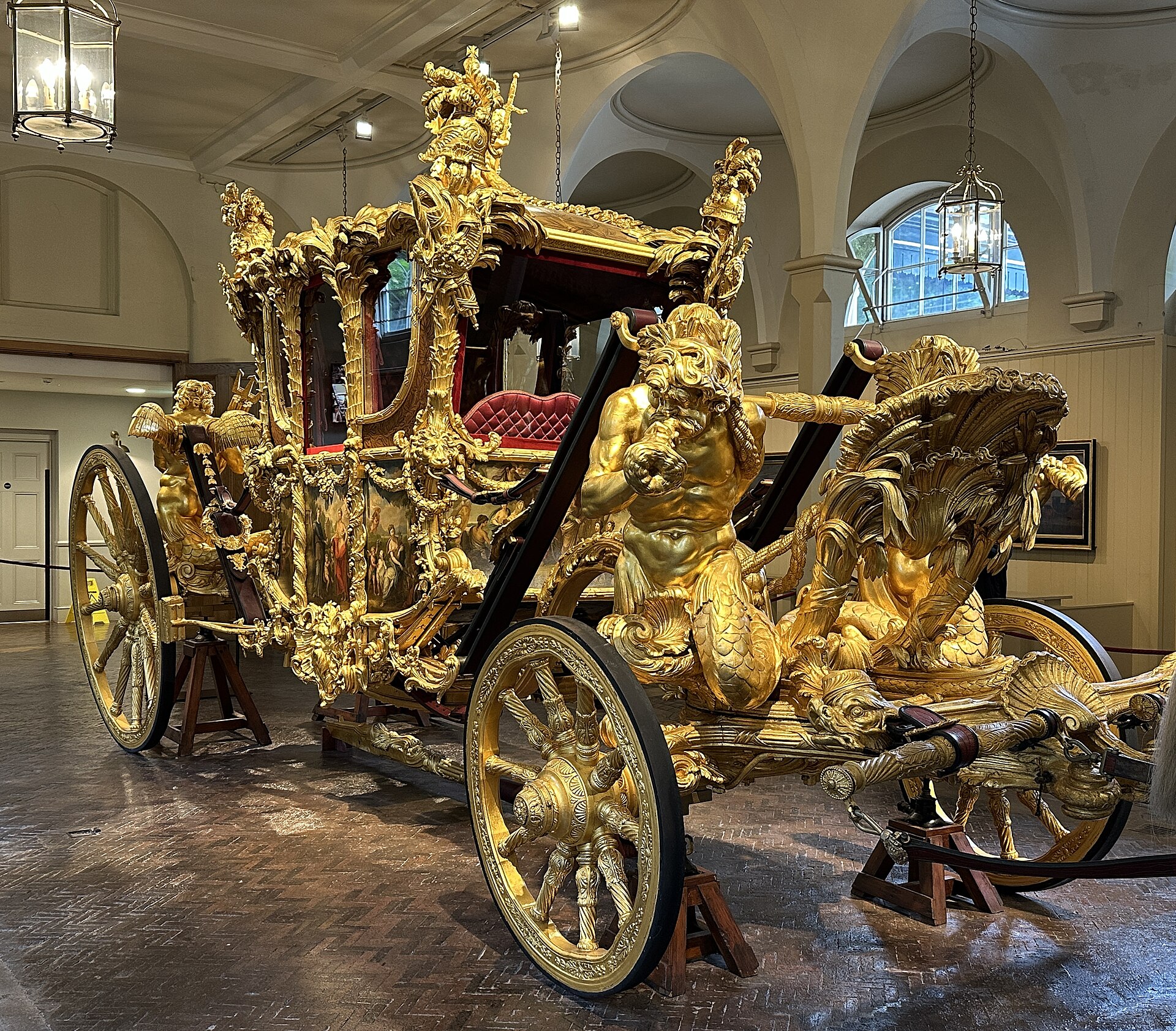 The Gold State Coach on display at the Royal Mews, covered in gilded carvings of tritons and palm trees, with painted panels on its sides and a red velvet seat inside