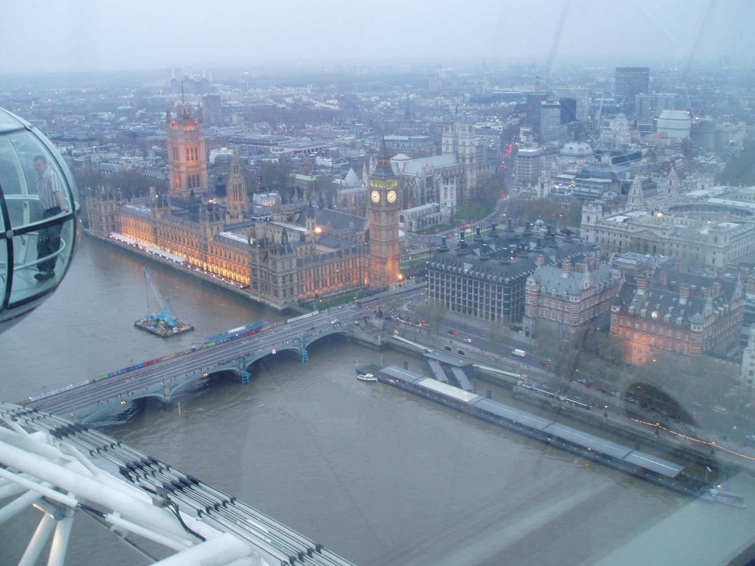 The Houses of Parliament and Big Ben seen from high up on the London Eye, with Westminster Bridge crossing the Thames below and the edge of a capsule on the left