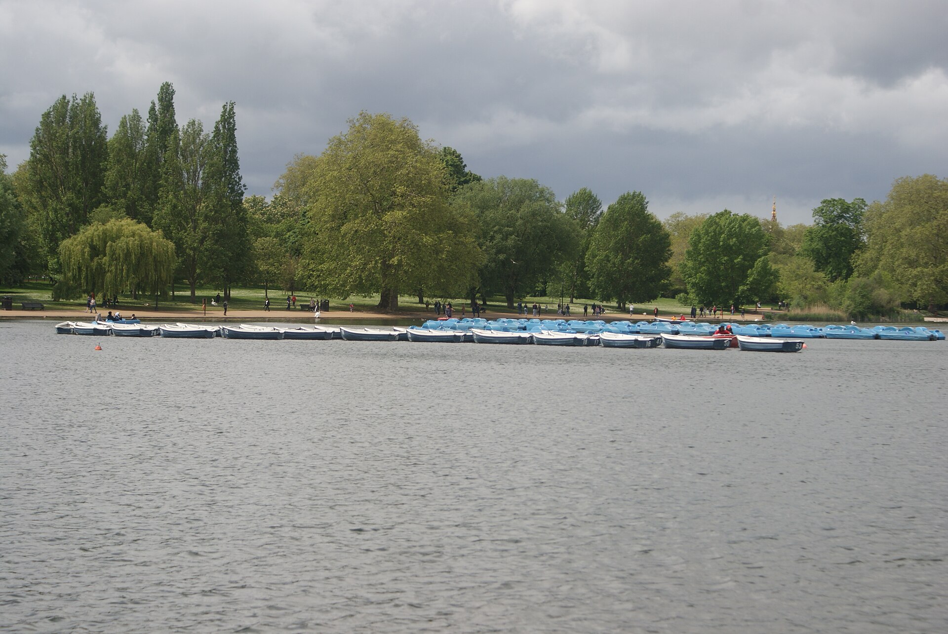 The Serpentine in Hyde Park with a row of blue hire boats moored along the far bank under a cloudy sky, tall trees behind