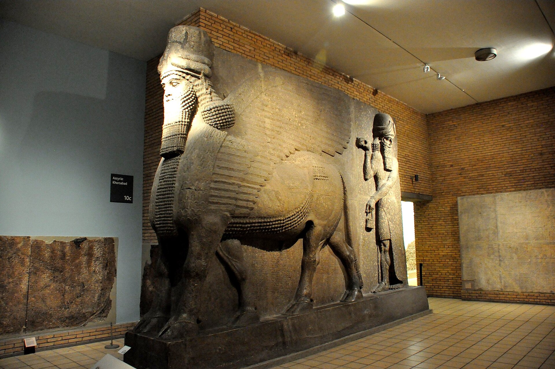 A human-headed winged bull with a curled beard from the palace of Sargon II at Khorsabad, carved from a huge stone block beside a winged divine figure, in Room 10 of the British Museum