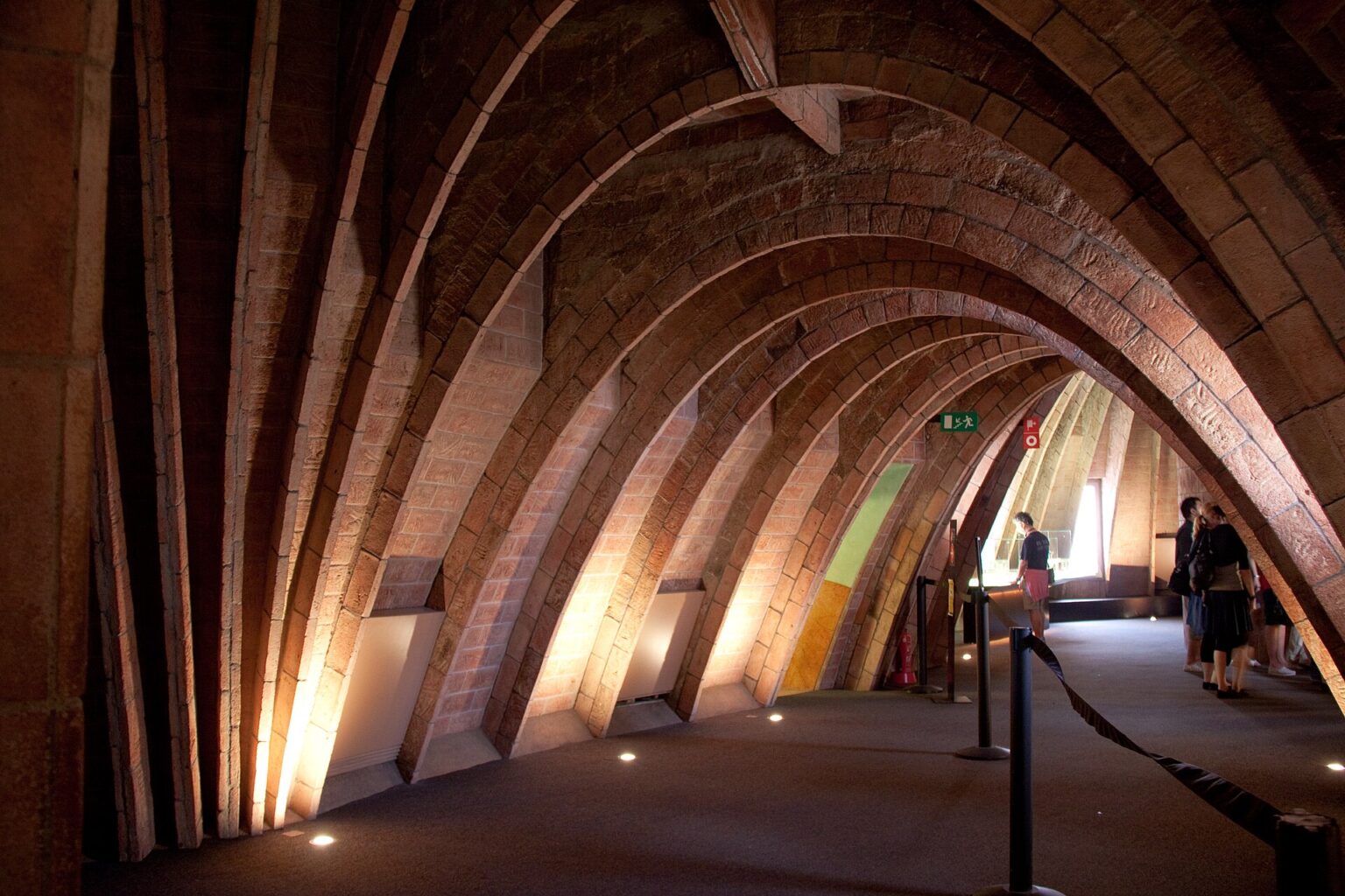 The attic of La Pedrera (Casa Milà): a long tunnel of bare brick catenary arches lit in warm orange, with visitors walking between the Gaudí exhibition displays