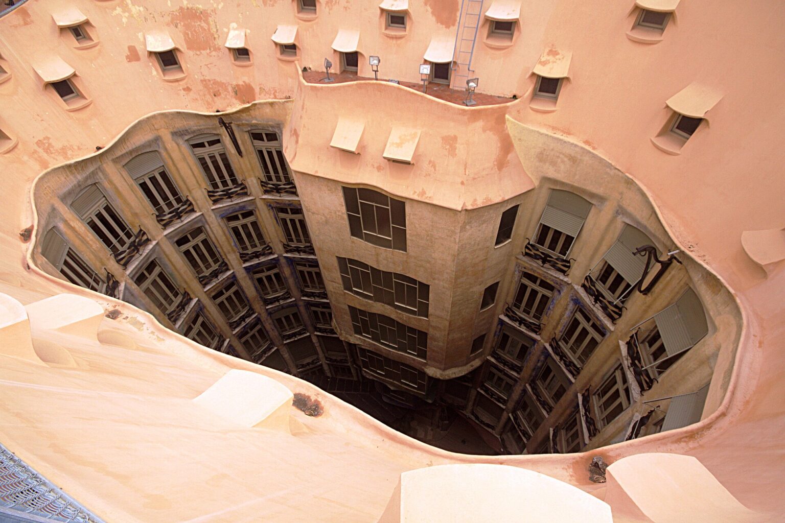 Looking down from the rooftop of La Pedrera (Casa Milà) into one of its 2 inner courtyards: an irregular oval opening ringed by small attic windows, with the flats' windows and balconies dropping down to the ground floor