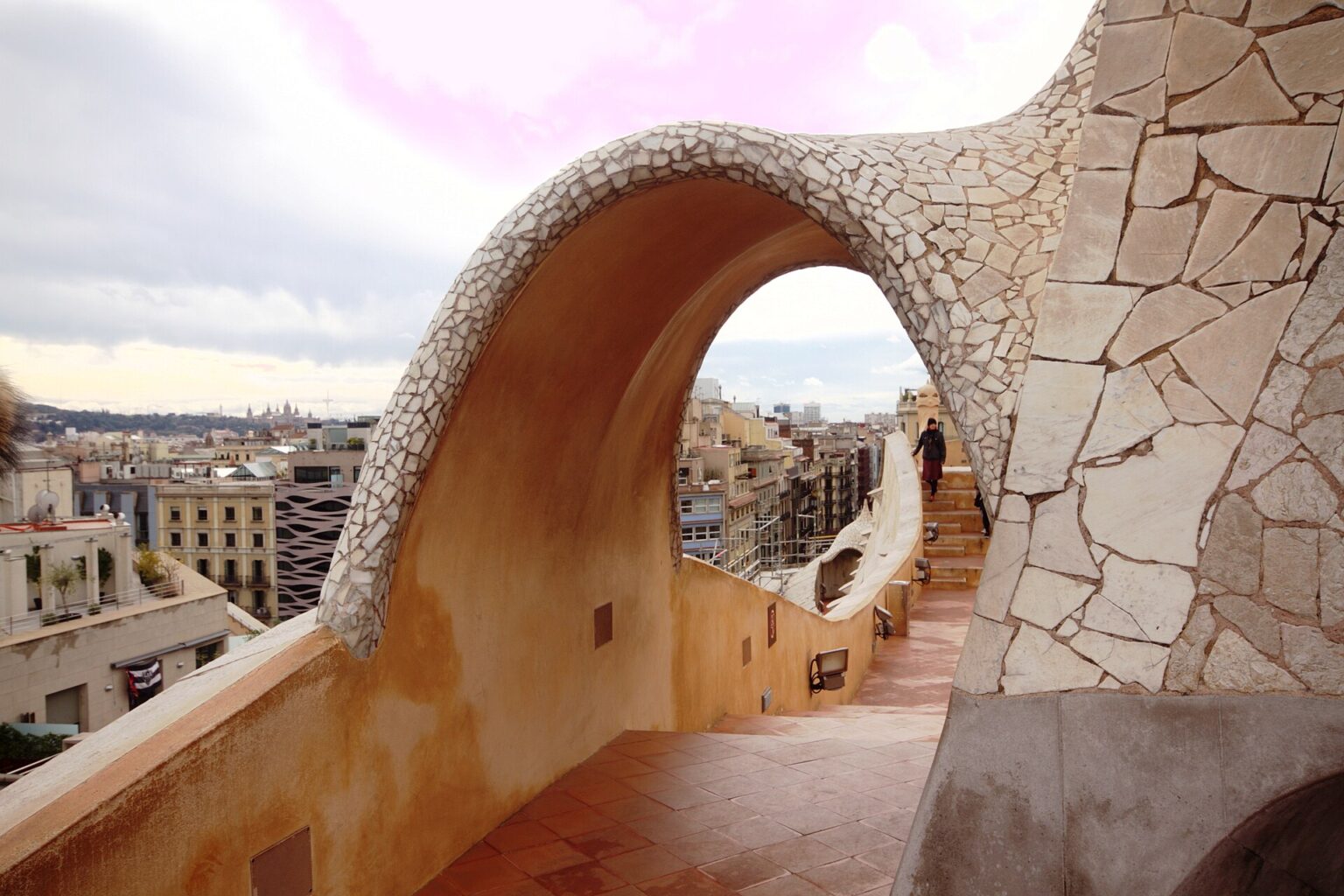 An arch clad in white broken-stone mosaic spanning a stairway on the rooftop of La Pedrera (Casa Milà), with the rooftops of the Eixample visible through it