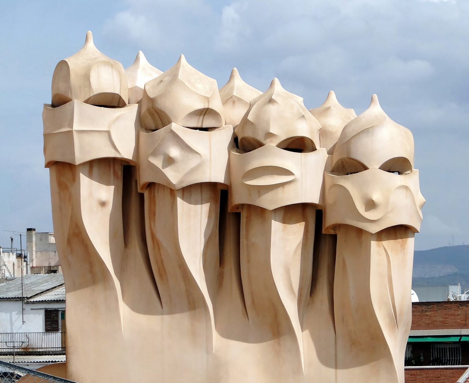 A cluster of 7 cream-coloured chimneys on the rooftop of La Pedrera (Casa Milà) in Barcelona, each shaped like a helmeted head with a visor, against a blue sky