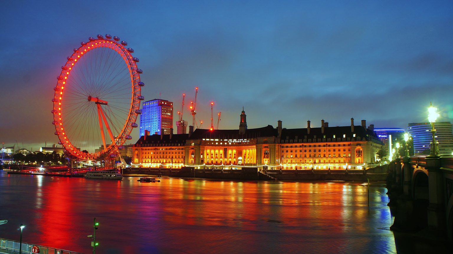 The London Eye lit red at dusk on the South Bank of the Thames, next to the lit-up County Hall building, seen from the end of Westminster Bridge