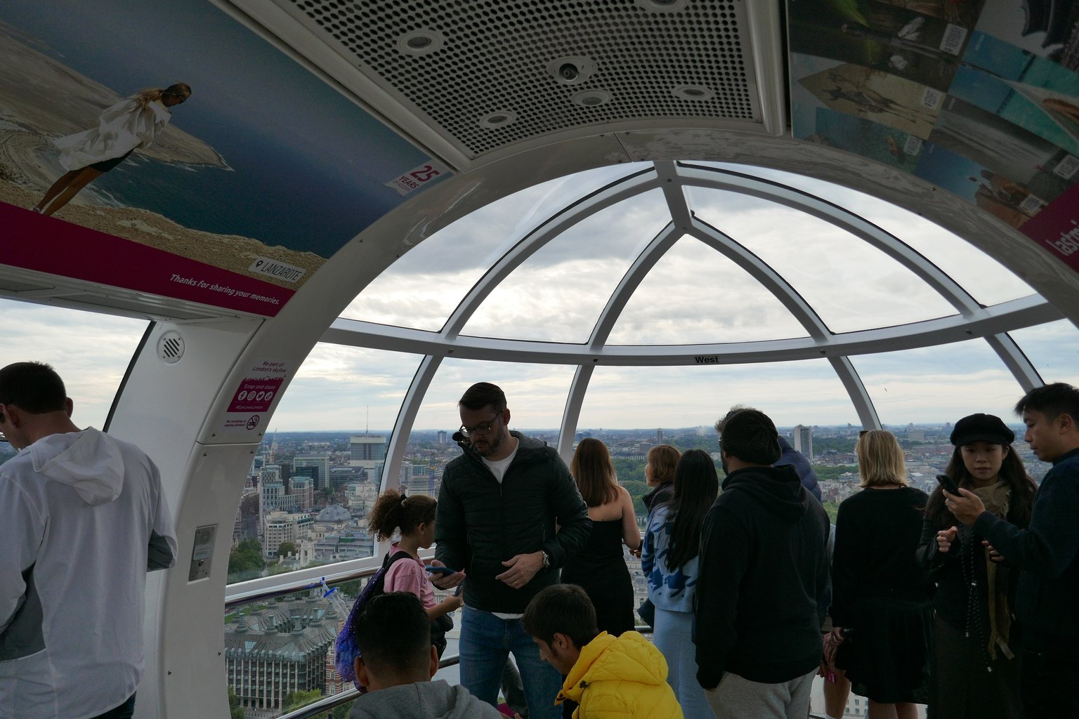 Inside a London Eye capsule: visitors standing at the curved glass and looking out over the rooftops of London on a cloudy day