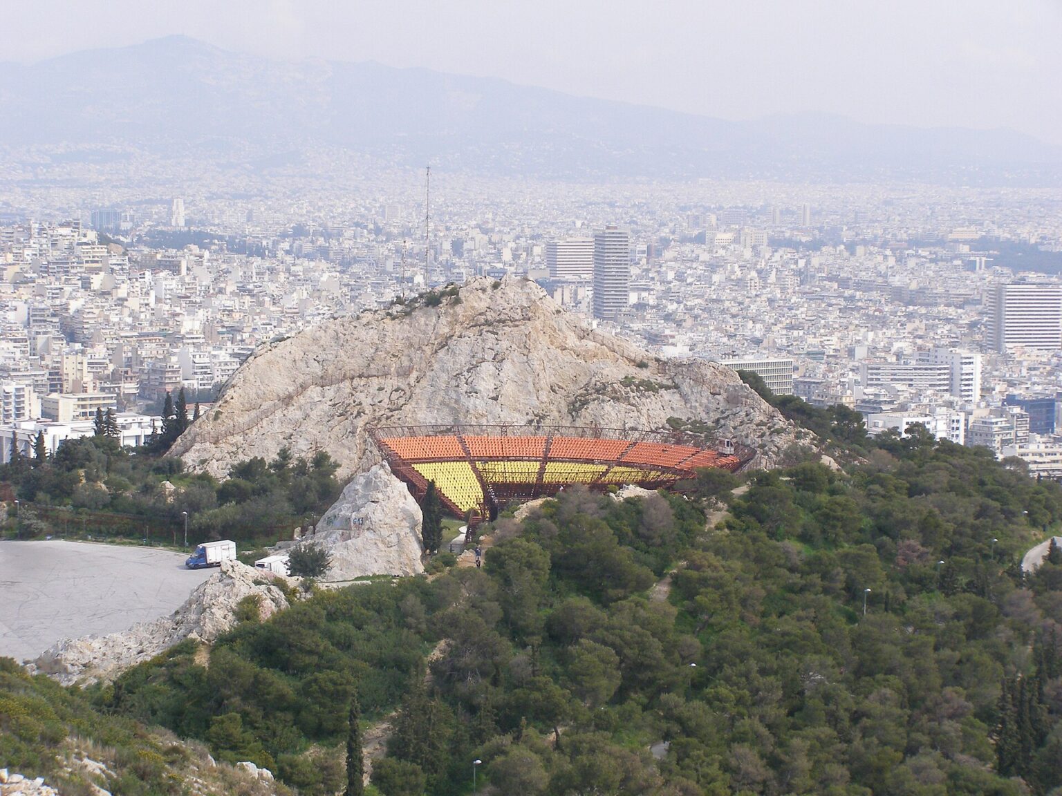 The open-air Lycabettus theatre seen from above, its yellow and orange seating tiers set into the hollow of an old quarry cut into the rock, with Athens spread out behind