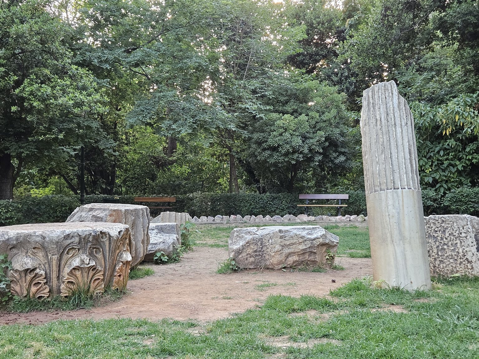 Ancient marble pieces on the grass of the National Garden of Athens: on the left a Corinthian capital carved with acanthus leaves, on the right a standing fluted column shaft, with benches and trees behind