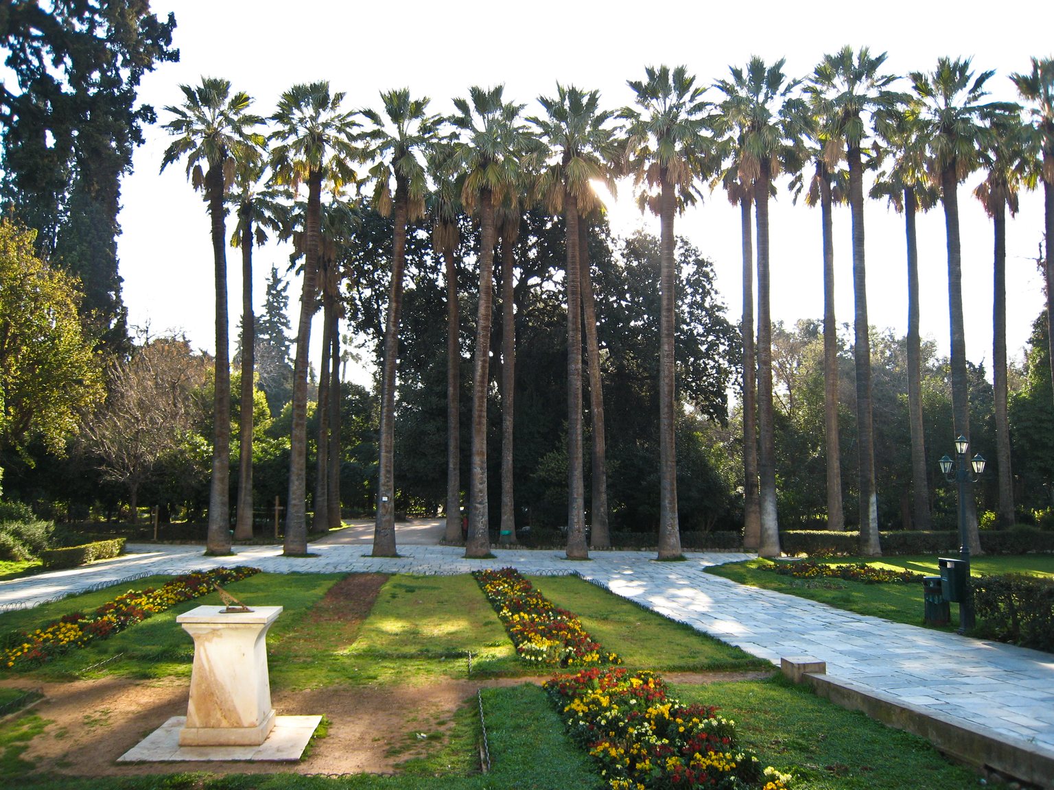 A row of very tall palm trees against the light in the National Garden of Athens, a path of marble slabs, beds of yellow and red flowers and, in the foreground, a sundial on a white marble pedestal