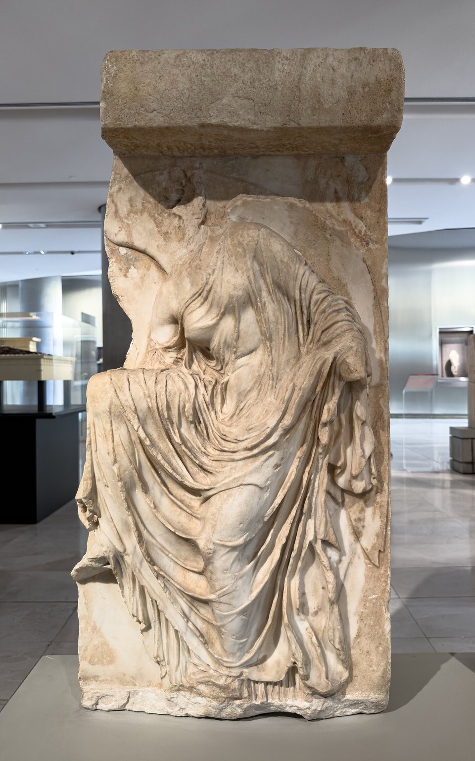 The marble slab of the Nike adjusting her sandal, standing in a gallery of the Acropolis Museum: the headless figure leans forward and her dress clings to her body in very fine folds that slip from her shoulder