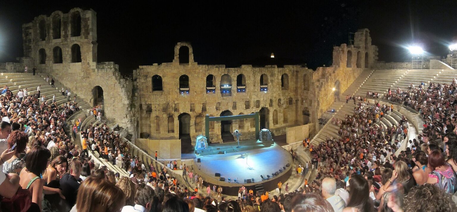 The Odeon of Herodes Atticus full for an evening performance: the audience fills every tier and floodlights mounted on the stage wall light the stage