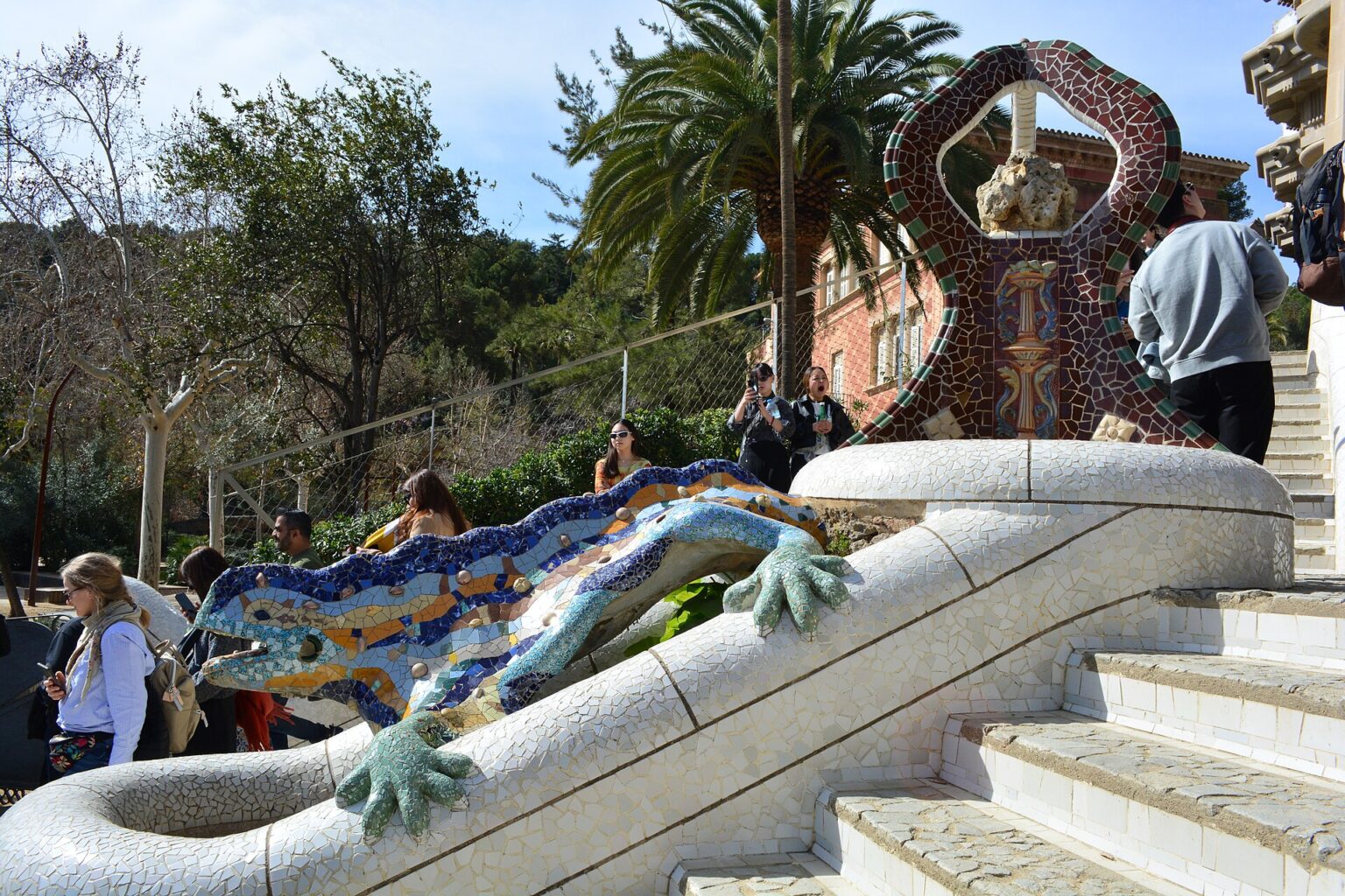 The mosaic dragon of Park Güell on the Dragon Stairway: a green, blue and orange tiled salamander stretched over white stone steps, with visitors taking photos and a red and white tiled pavilion roof behind