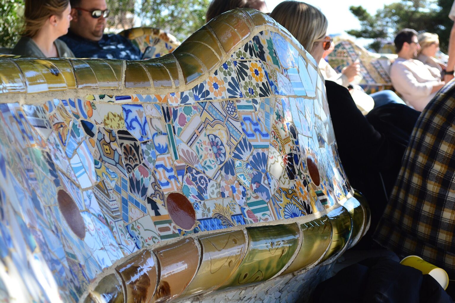 Close-up of the undulating bench on Nature Square in Park Güell, covered in a mosaic of blue, white, yellow and green ceramic fragments, with visitors sitting along it in the sun