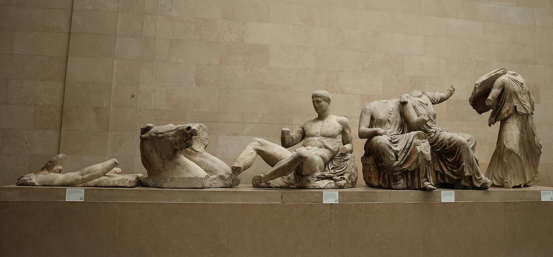 Marble figures from a pediment of the Parthenon displayed in a row on a plinth in Room 18 of the British Museum: a horse's head, a reclining youth and three headless female figures