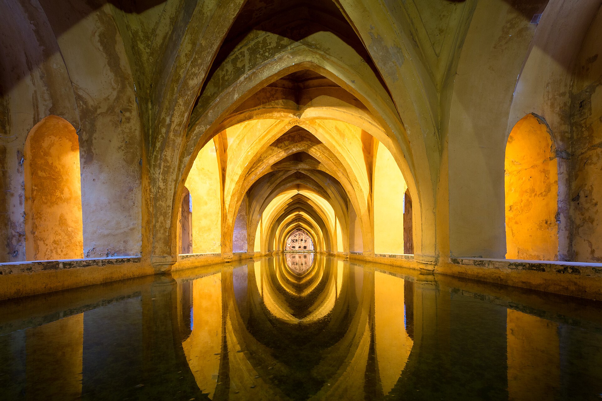 The Baths of María de Padilla under the Real Alcázar of Seville: a long vaulted gallery of pointed arches lit in warm yellow, reflected in the still, dark water of the pool that fills its floor