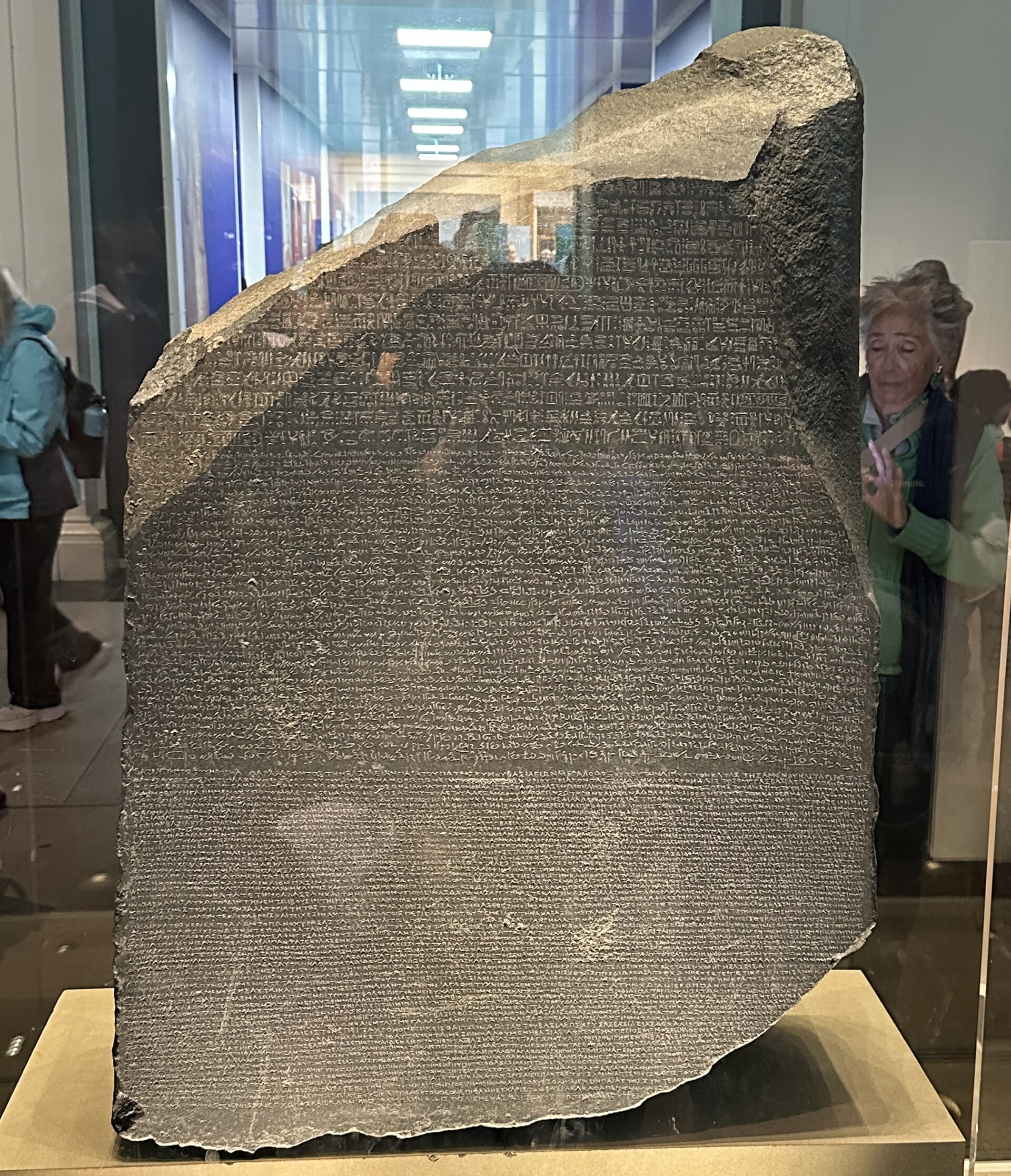 The Rosetta Stone, a dark stone slab with a broken top corner and its face covered in lines of carved text, in its glass case in Room 4 of the British Museum with visitors behind