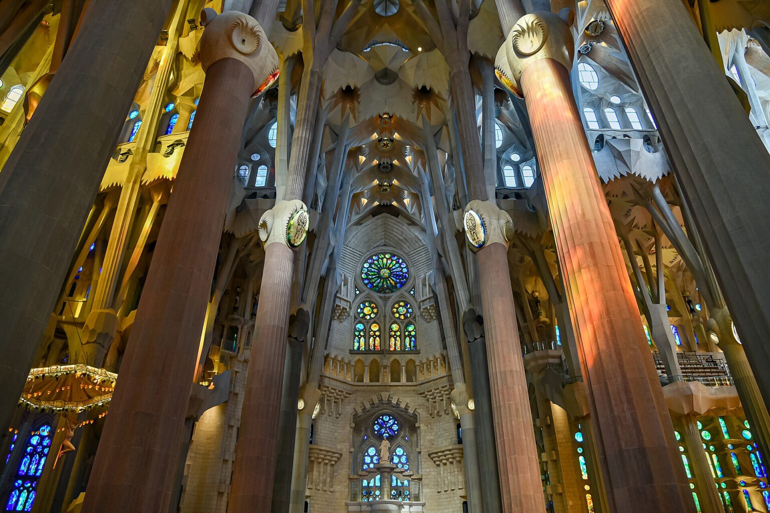 Main nave of the Sagrada Familia looking towards the apse: tall tree-like stone columns branching into the vaults, sunlight through red and yellow stained glass on the left and blue and green windows around the apse