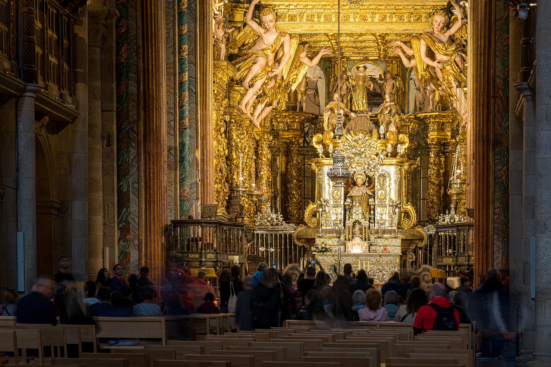 The gilded baroque high altar of Santiago Cathedral with angels on the canopy, and visitors sitting in the pews of the nave