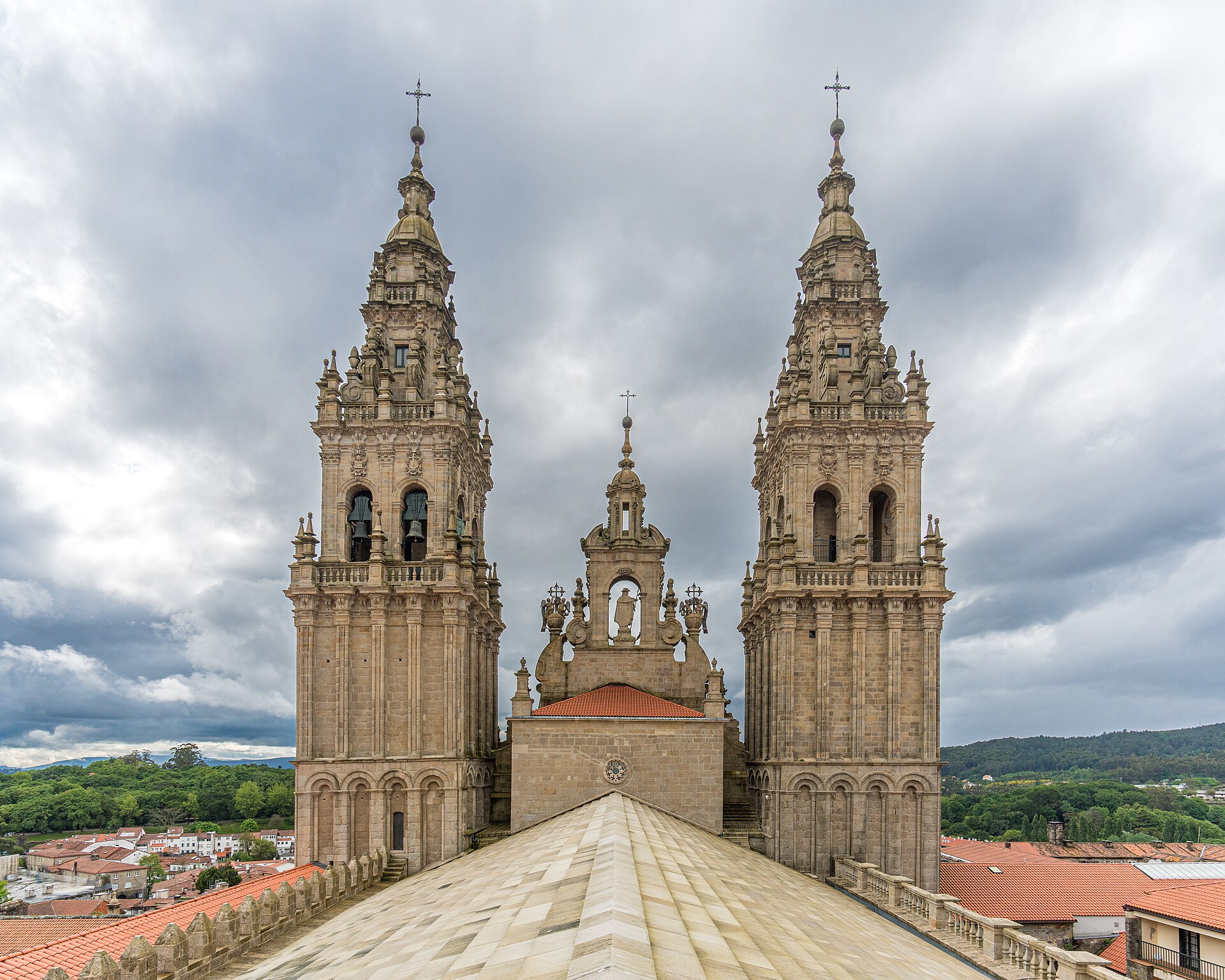 The stepped granite rooftops of Santiago Cathedral with the two towers of the Obradoiro facade ahead, under a cloudy sky