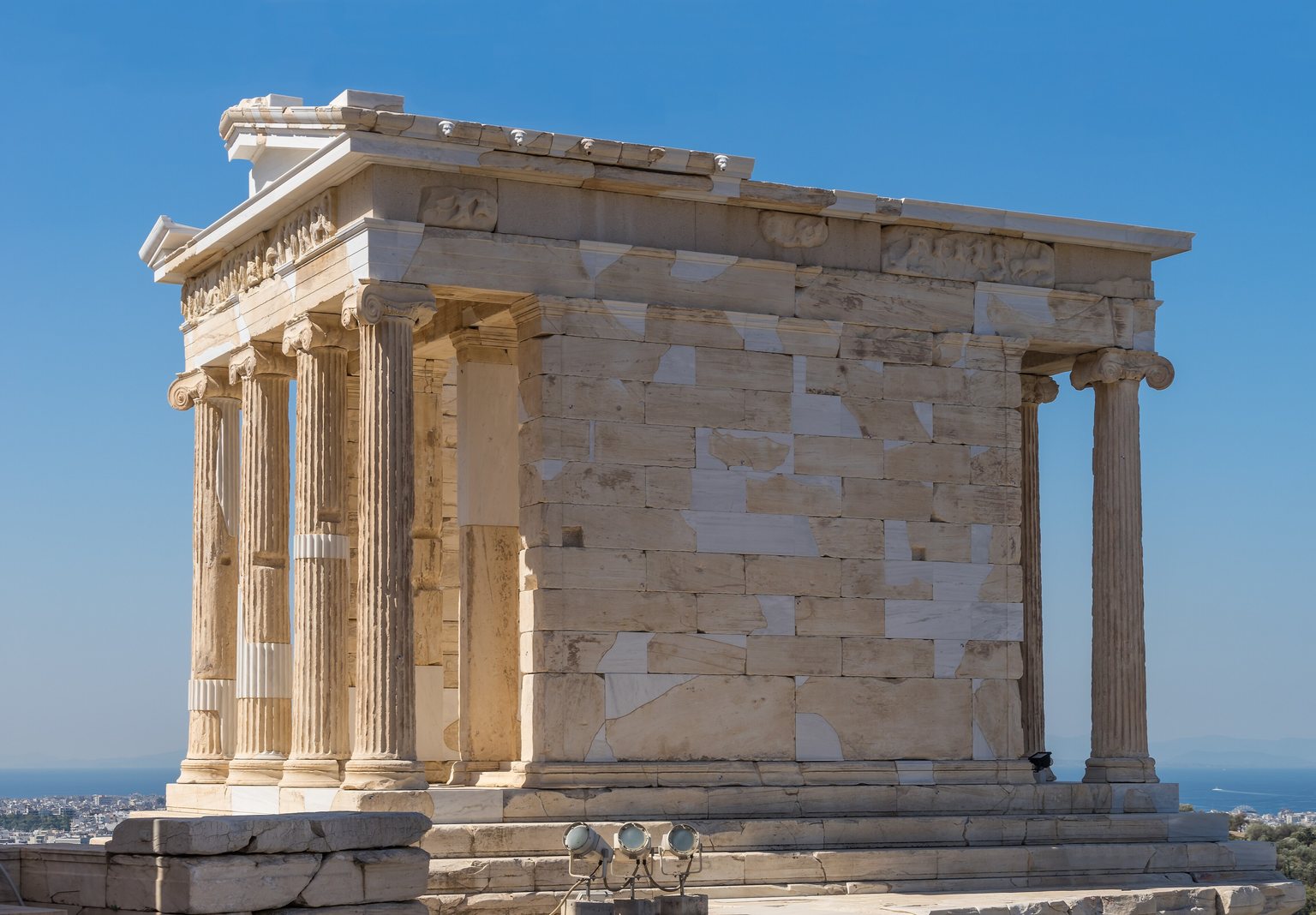 The Temple of Athena Nike seen from one corner: the 4 Ionic columns at one end, the plain wall of the cella with no columns down the side, and the columns of the far end behind, under a clear sky with the sea on the horizon