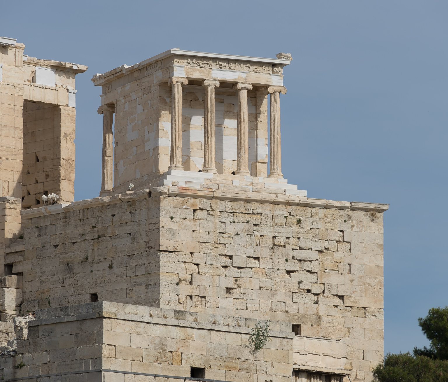 The Temple of Athena Nike perched on top of its bastion, seen from below: the wall of squared blocks fills half the picture and the much smaller temple rises above it, with the wall of the Propylaia on the left