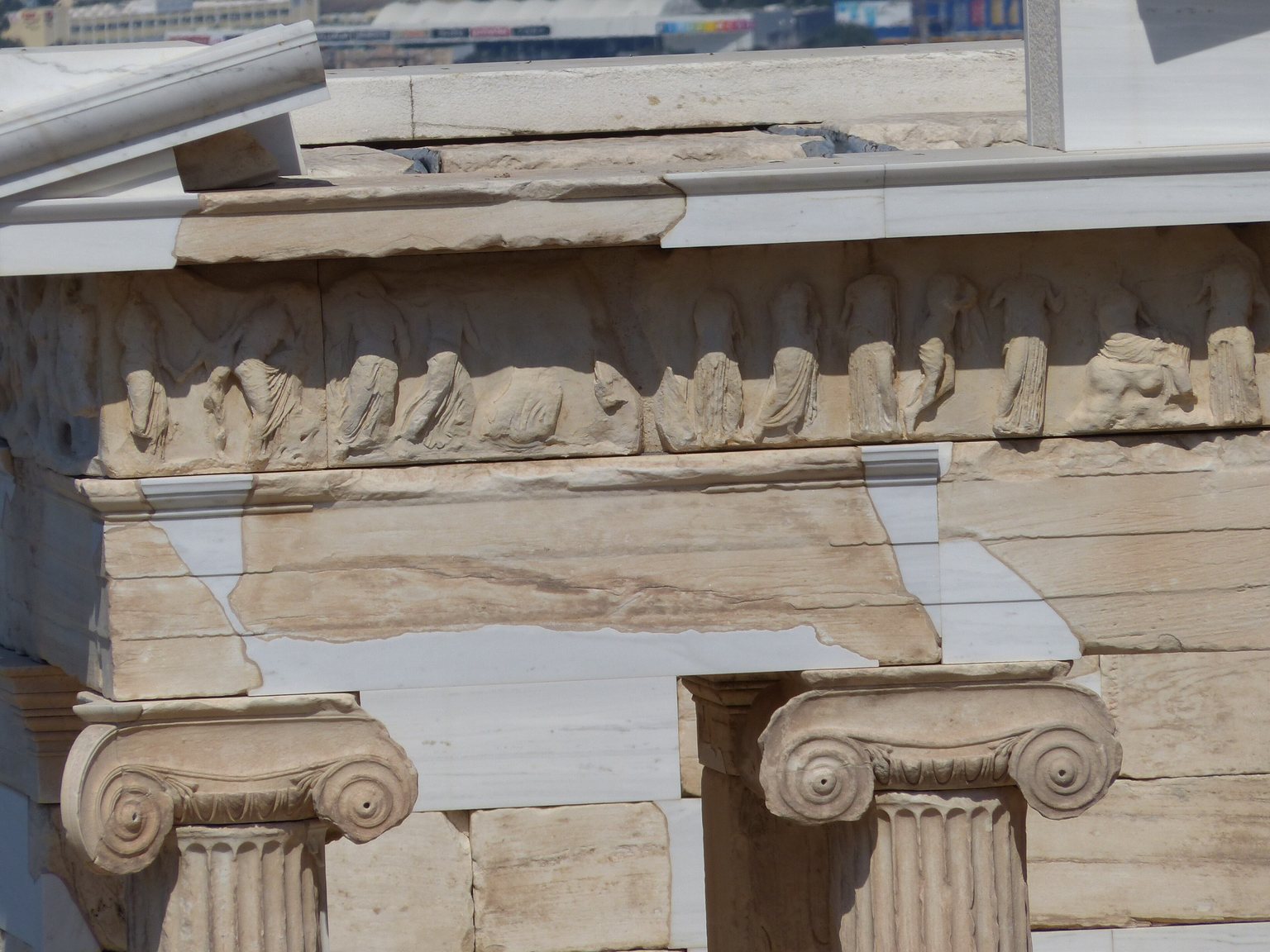 Close view of the frieze of the Temple of Athena Nike: a narrow band of figures in deeply folded robes, one of them seated and a horse at the right-hand end, above the architrave and 2 Ionic capitals with their volutes