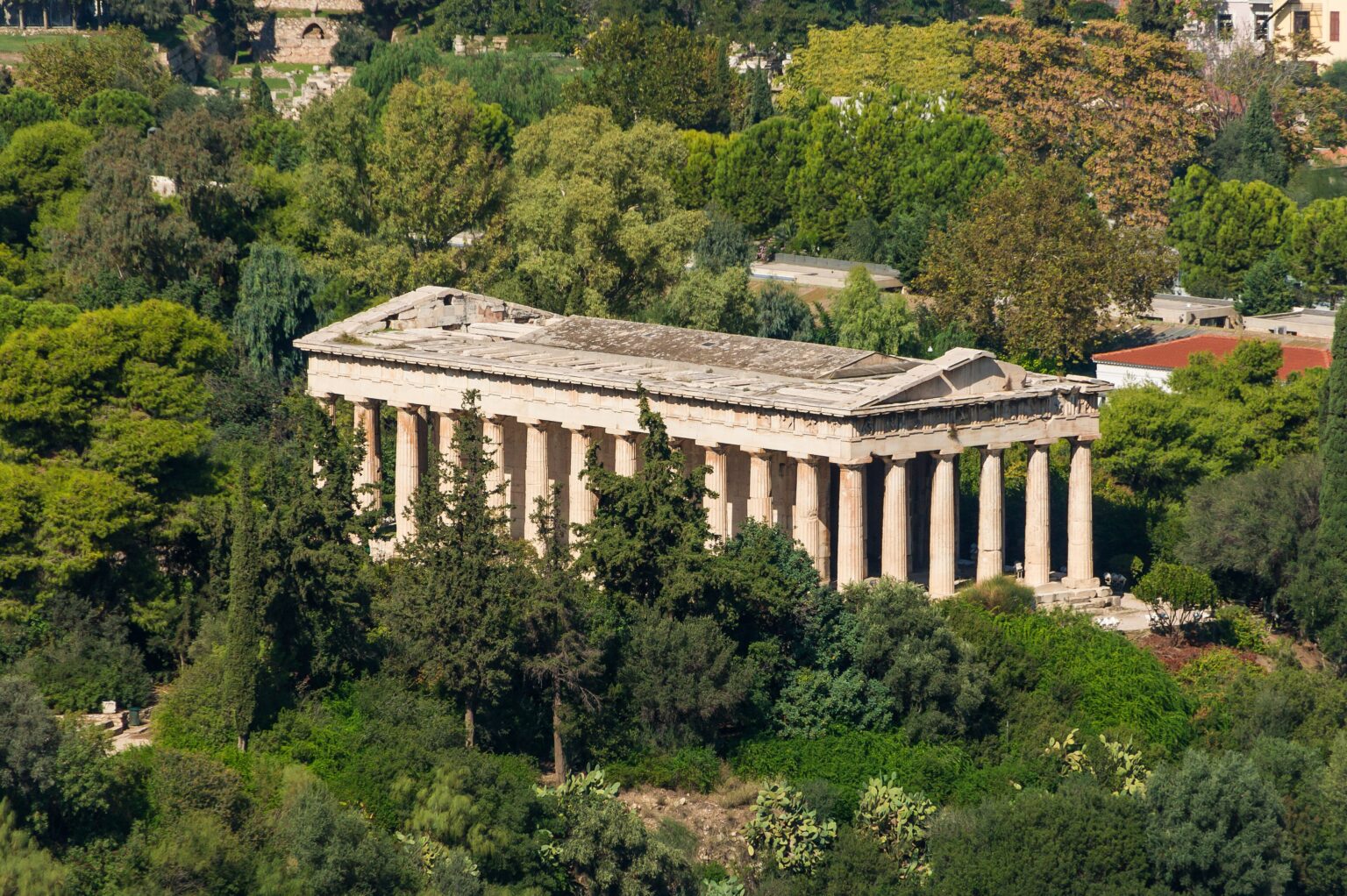 The Temple of Hephaestus seen from the top of the Acropolis, whole and side on above its low hill, ringed by the pines and cypresses of the Ancient Agora with rooftops of the neighbourhood behind on the right