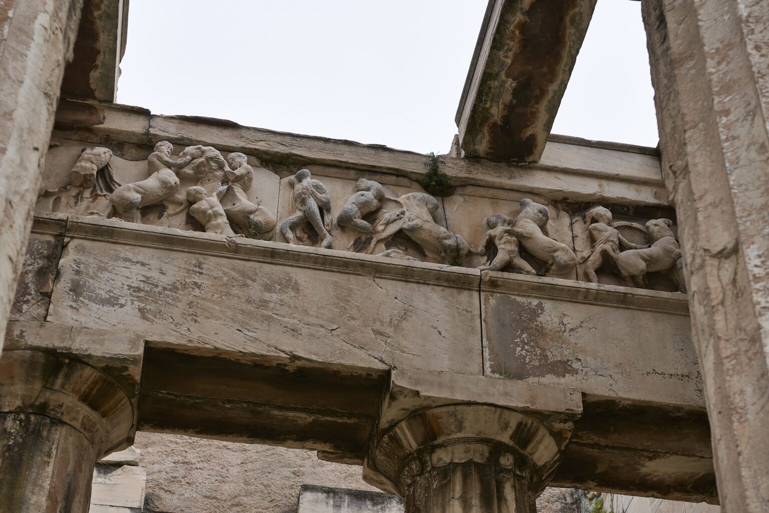 Close view from below of the carved frieze of the Temple of Hephaestus: naked human figures wrestling hand to hand with centaurs, in high relief on the marble band, with two Doric capitals underneath