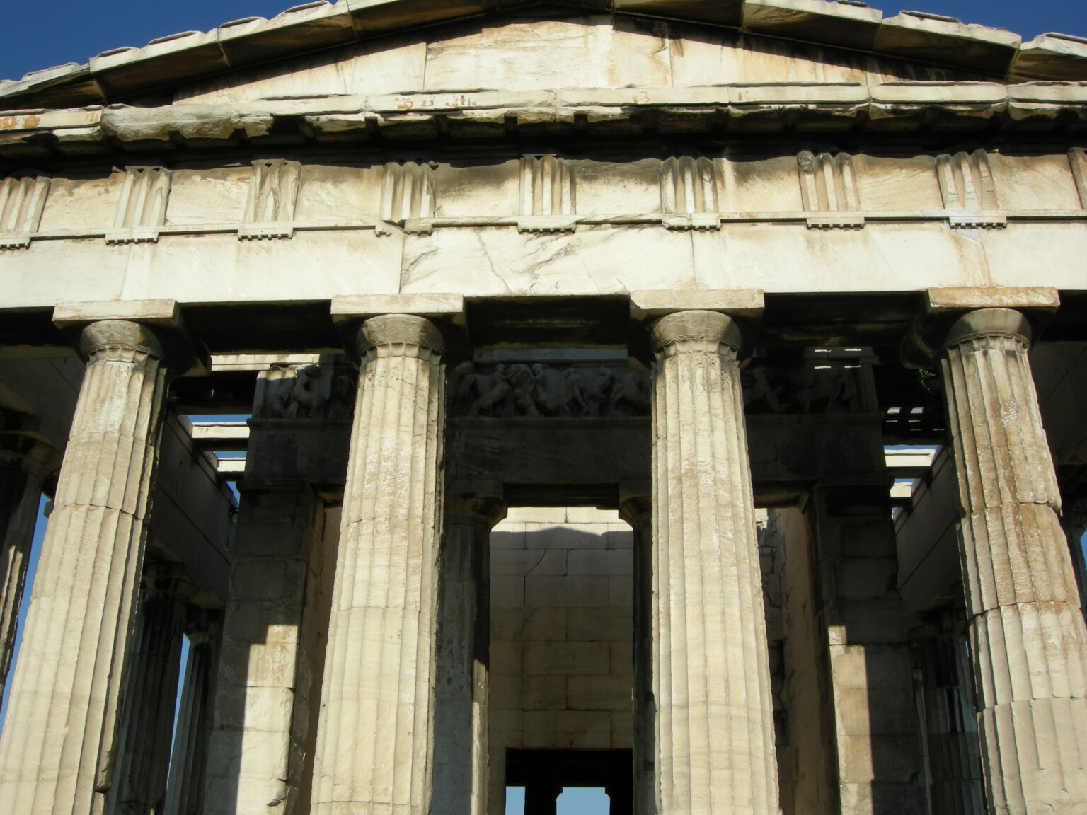The front of the Temple of Hephaestus seen head on from below, with the empty triangular pediment, the row of triglyphs and metopes across the entablature and the fluted Doric columns; the carved frieze shows in the shadow behind them