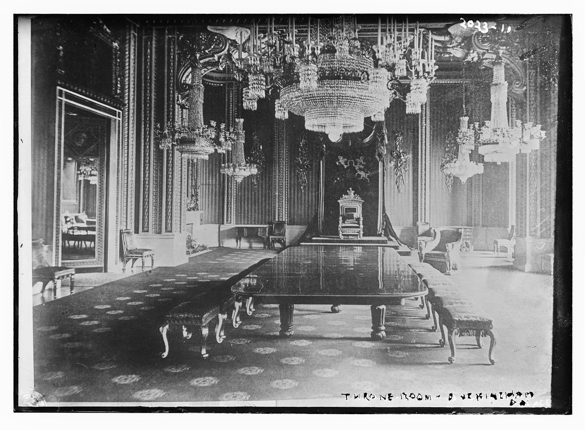 Old black-and-white photograph of the Throne Room at Buckingham Palace, with crystal chandeliers, a long polished table and the throne on a dais under a canopy