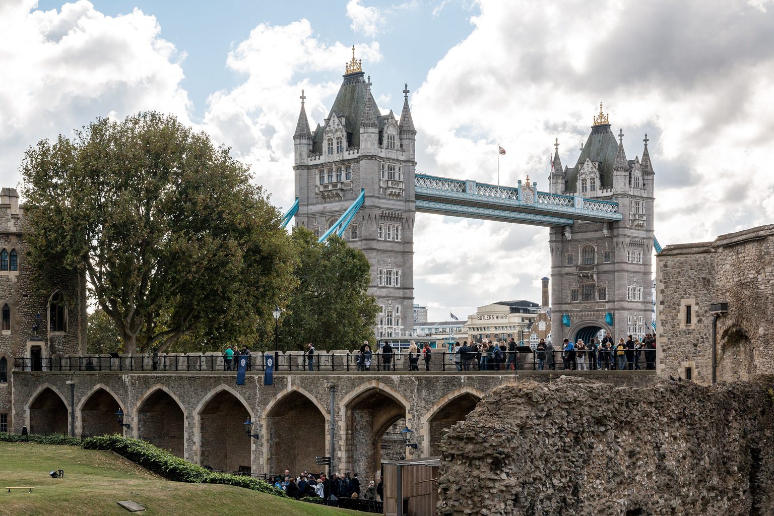 The tops of the 2 towers of Tower Bridge rising behind the stone walls and arches of the Tower of London, with trees, a lawn and visitors walking along the wall