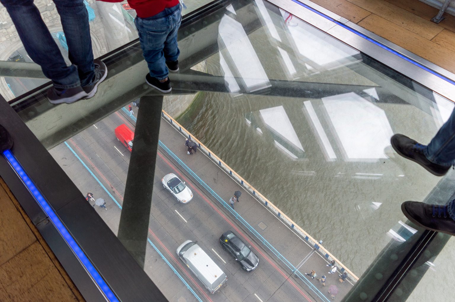 Looking down through the glass floor of a Tower Bridge walkway: visitors' feet on the glass and, far below, cars and people on the road deck and the water of the Thames