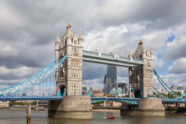Tower Bridge seen across the River Thames on a cloudy day: the 2 stone towers joined by the high-level walkways, the suspension chains painted pale blue and a small red boat passing underneath