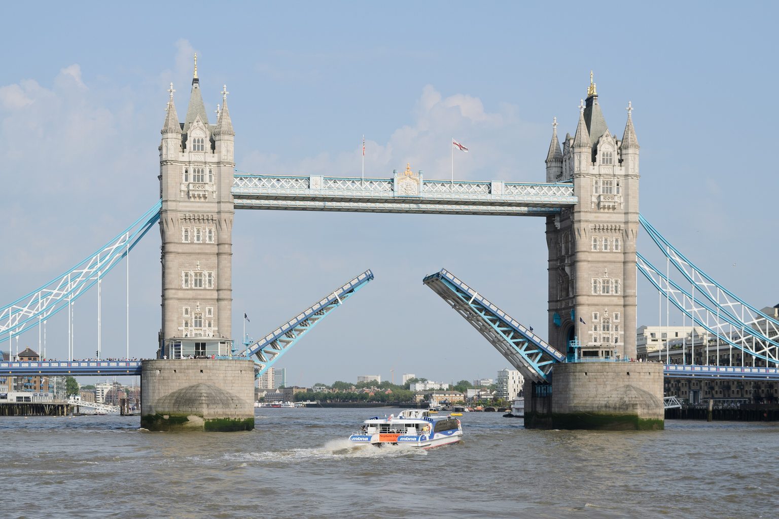 Tower Bridge open on a sunny day: both halves of the roadway raised at an angle between the towers while a white and blue boat sails through on the Thames