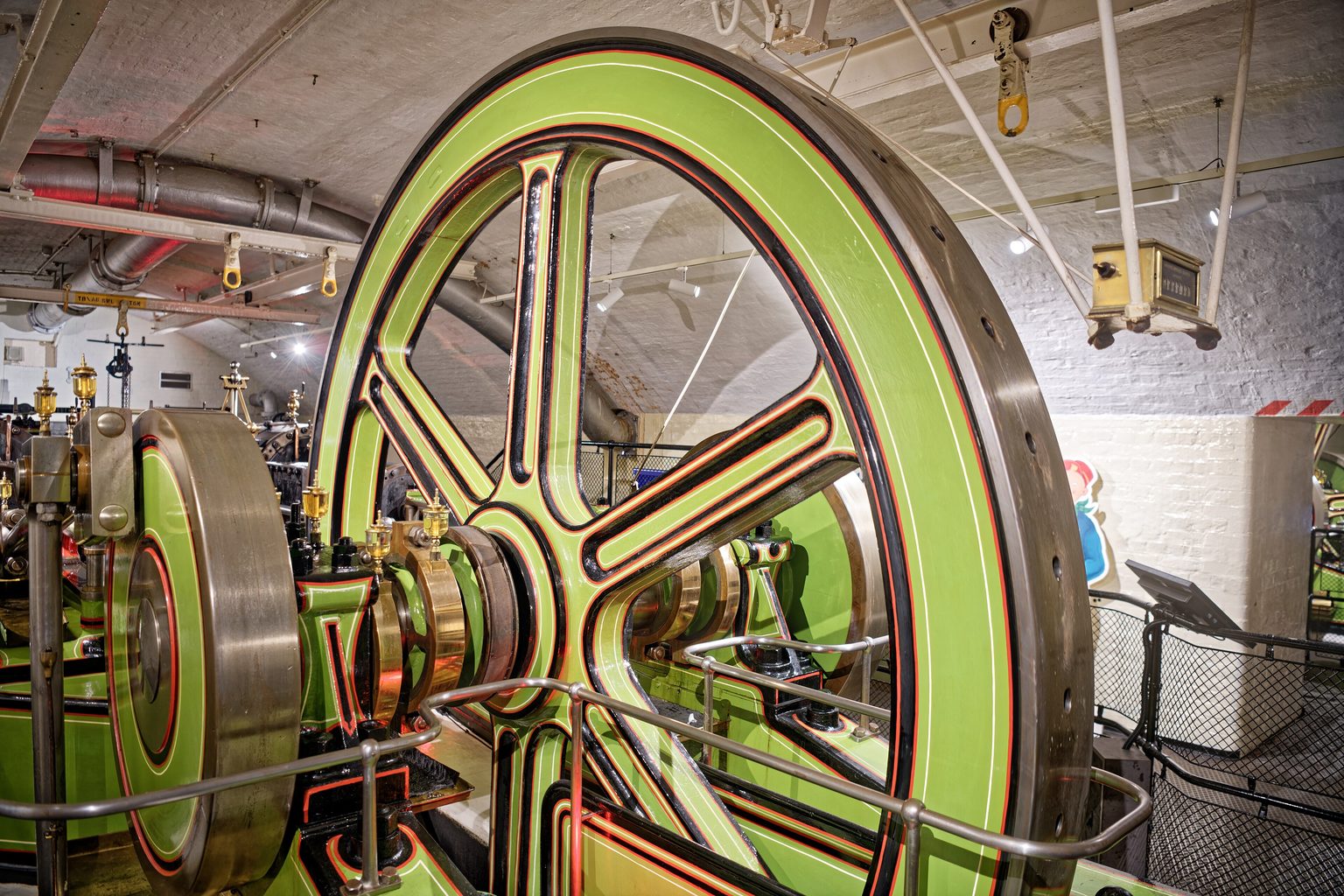 The large flywheel of a steam engine painted green with red and black lining in the Victorian Engine Rooms of Tower Bridge, under a vaulted ceiling of white brick with pipes