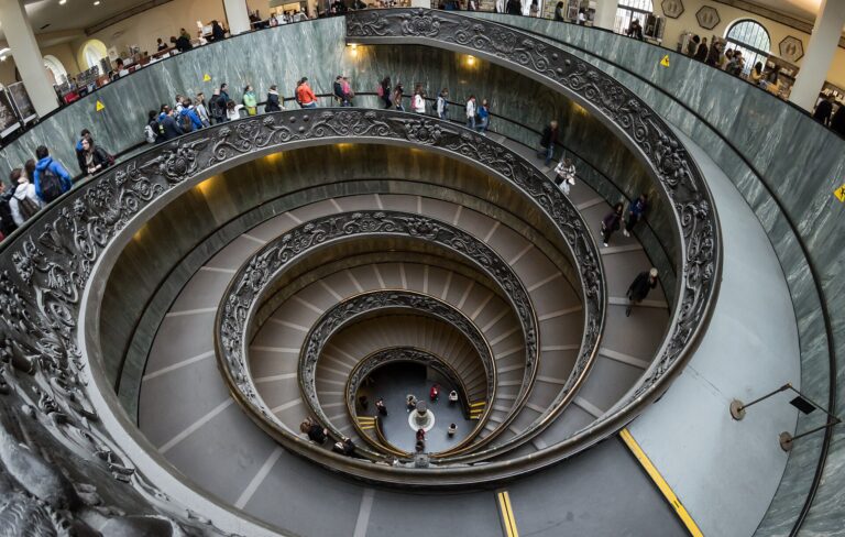 The double-helix spiral staircase at the exit of the Vatican Museums seen from above, with visitors walking down the bronze ramp