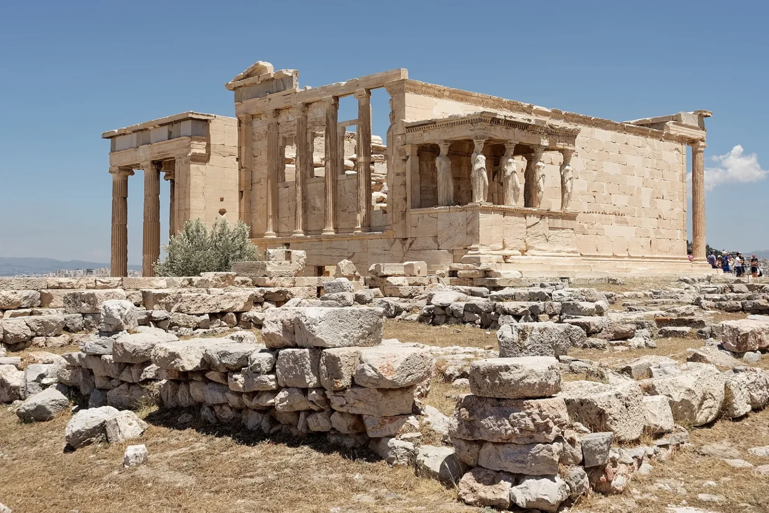 The Erechtheion seen from the south-west behind a field of fallen blocks: the north porch with its Ionic columns on the left, the west front in the middle and, on the right, the porch of the Caryatids, with figures of women instead of columns holding up the roof
