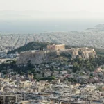 The Acropolis rock seen from Lycabettus Hill, rising out of the rooftops of Athens with the Parthenon on top, and the sea and the coast in the haze behind