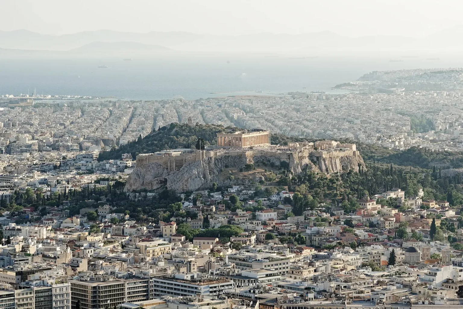 The Acropolis rock seen from Lycabettus Hill, rising out of the rooftops of Athens with the Parthenon on top, and the sea and the coast in the haze behind