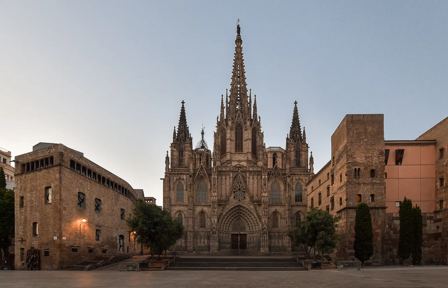 The neo-Gothic main facade of Barcelona Cathedral at dawn, with its tall central spire, 2 side towers and the wide stone steps of Pla de la Seu in front