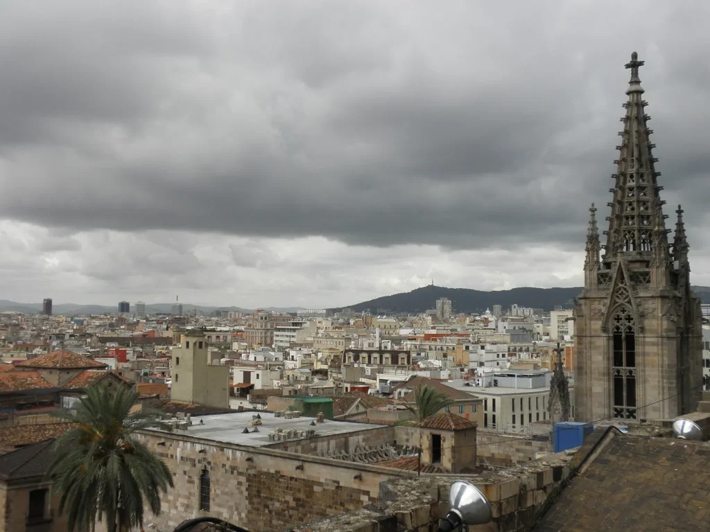 View from the rooftop of Barcelona Cathedral over the roofs of the old town towards the hills, with one of the cathedral's openwork Gothic spires on the right