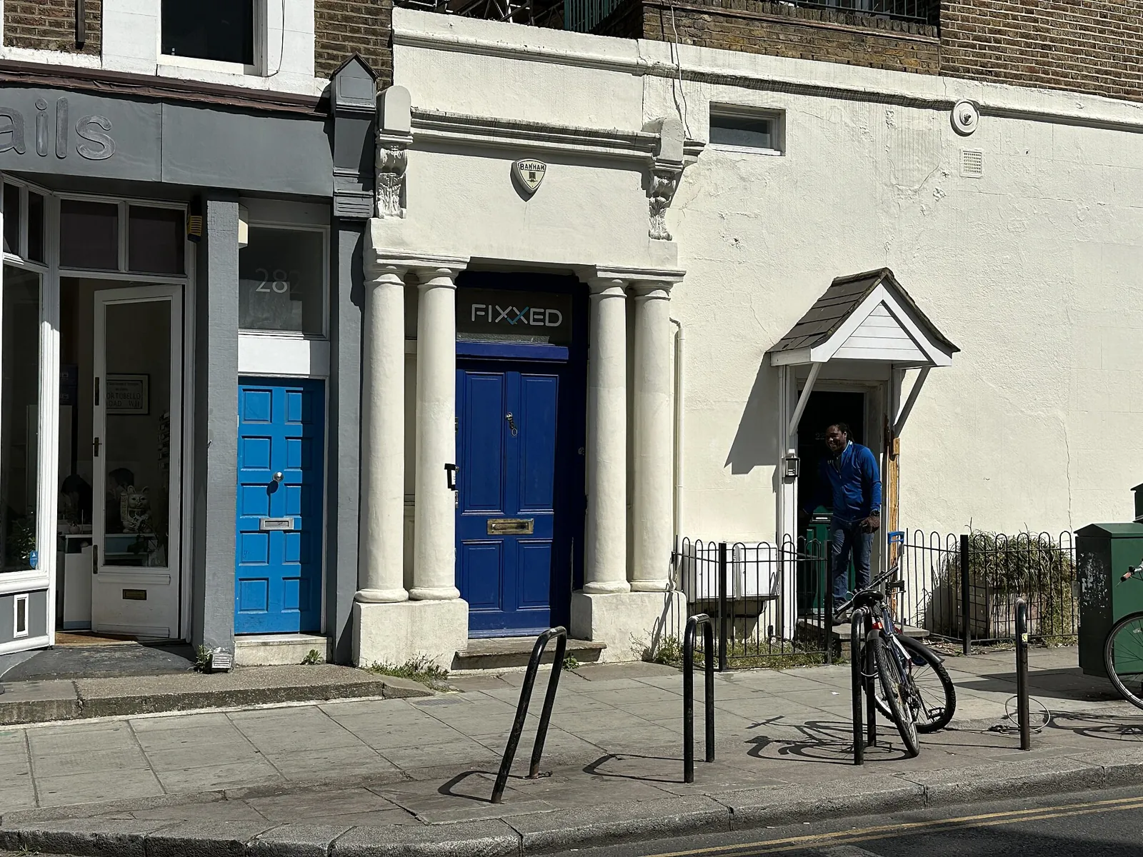 A white front with columns and a blue door on Westbourne Park Road, next to a smaller blue door and a bike rack