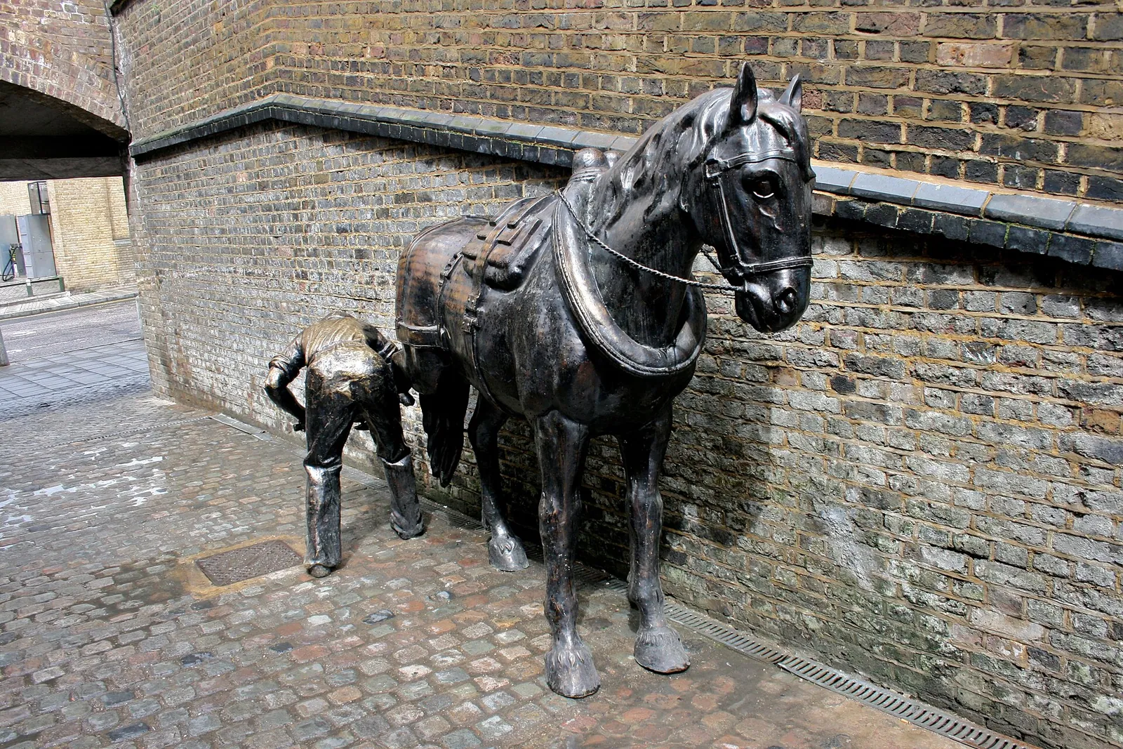Bronze sculpture of a harnessed draught horse and a farrier holding its hoof, by a brick wall in the Stables Market